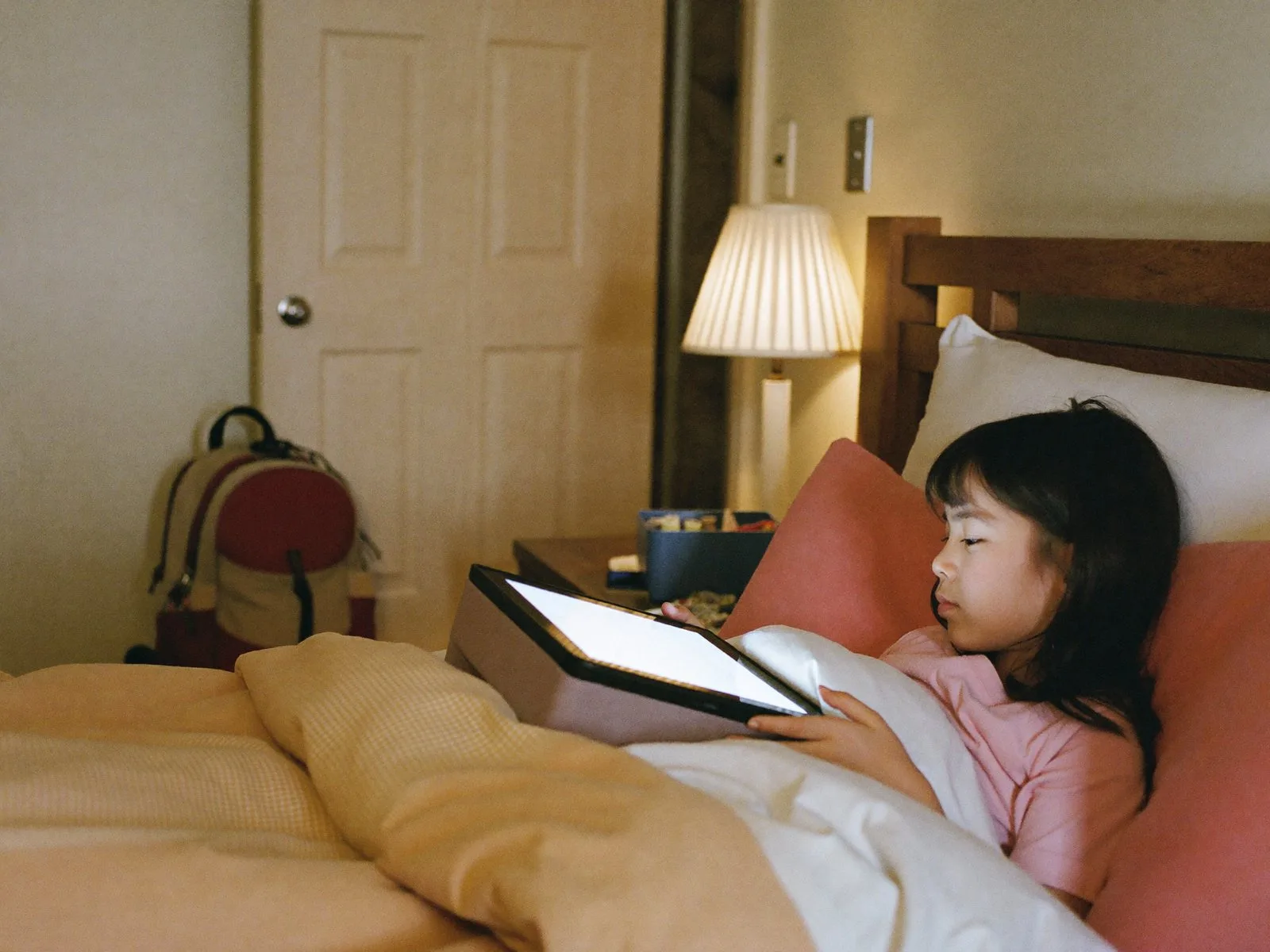 A child in bed at a second home listening to a story, an overnight bag by the door